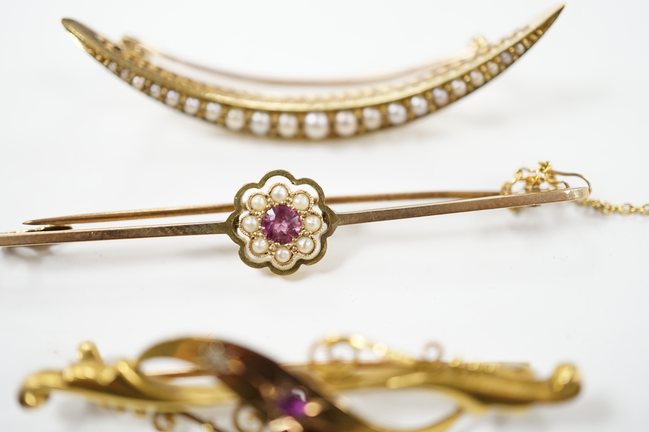 An Edwardian 15ct, ruby and diamond chip set three stone bar brooch, 45mm, a 9ct and gem set bar brooch and a yellow metal and seed pearl set crescent brooch.